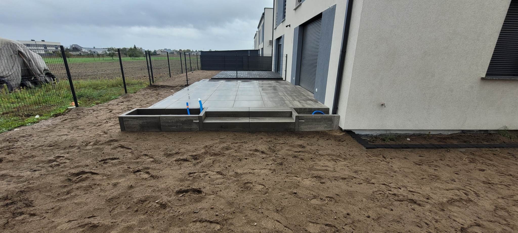 Nowoczesny taras z szarych płyt betonowych, z niską donicą i schodami, widoczne rury instalacji wodnej, w tle ogrodzenie panelowe i budynki mieszkalne.