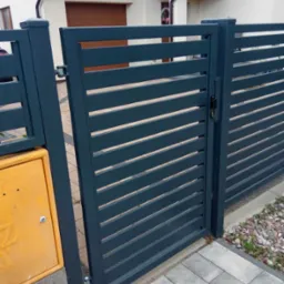 Nowoczesna, pozioma brama palisadowa w kolorze antracytowym, z widocznym zamkiem i zawiasami, zamontowana przy posesji z żółtą skrzynką gazową.