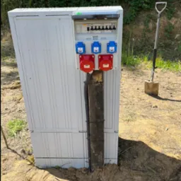 Biała skrzynka elektryczna z gniazdami siłowymi (czerwone i niebieskie) i zabezpieczeniami, umieszczona na drewnianym słupku, obok szpadel wbity w ziemię, na tle zieleni.