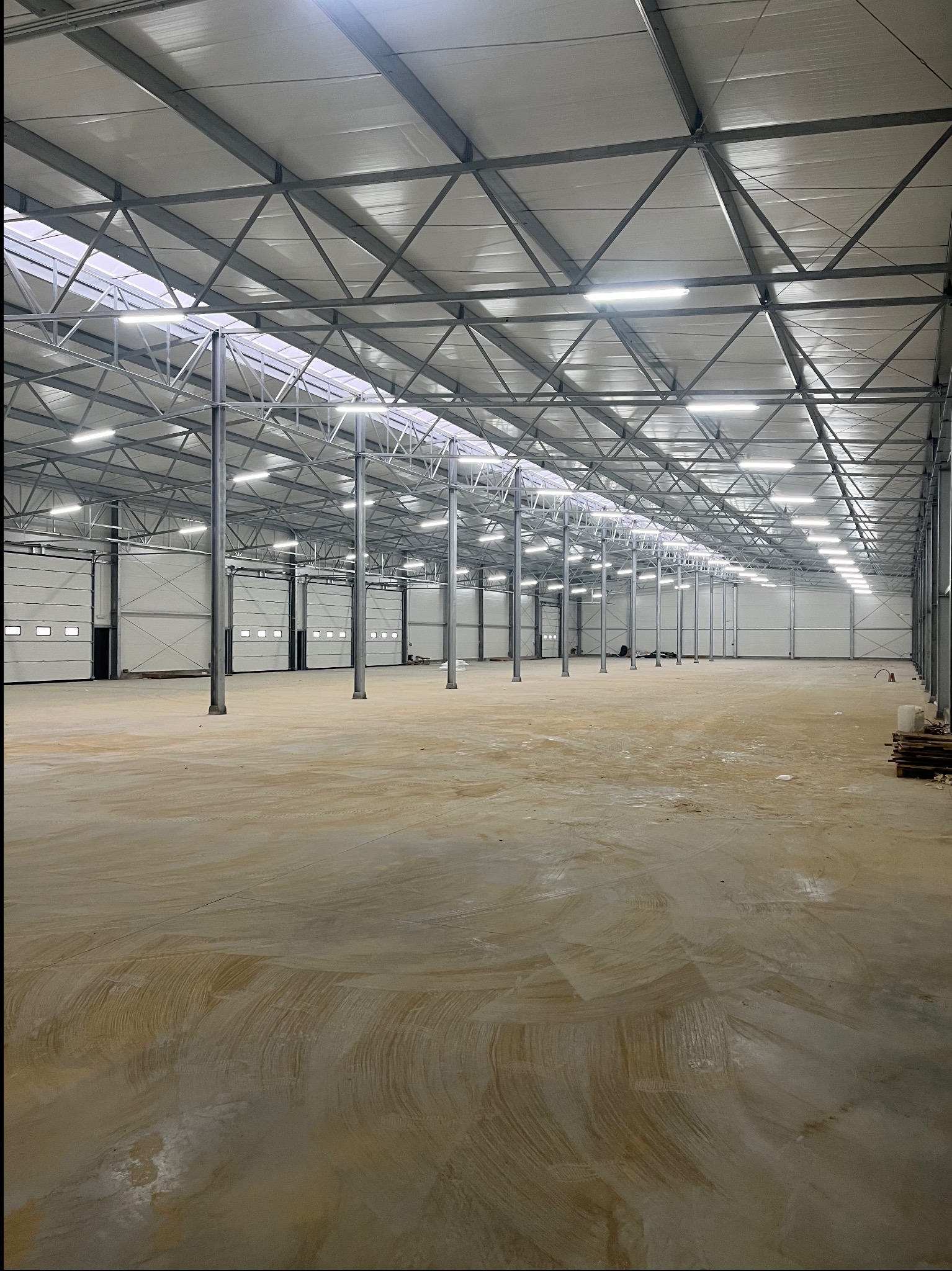 Wnętrze nowo wybudowanej hali magazynowej z betonową posadzką, stalową konstrukcją dachu z oświetleniem LED i bramami segmentowymi.