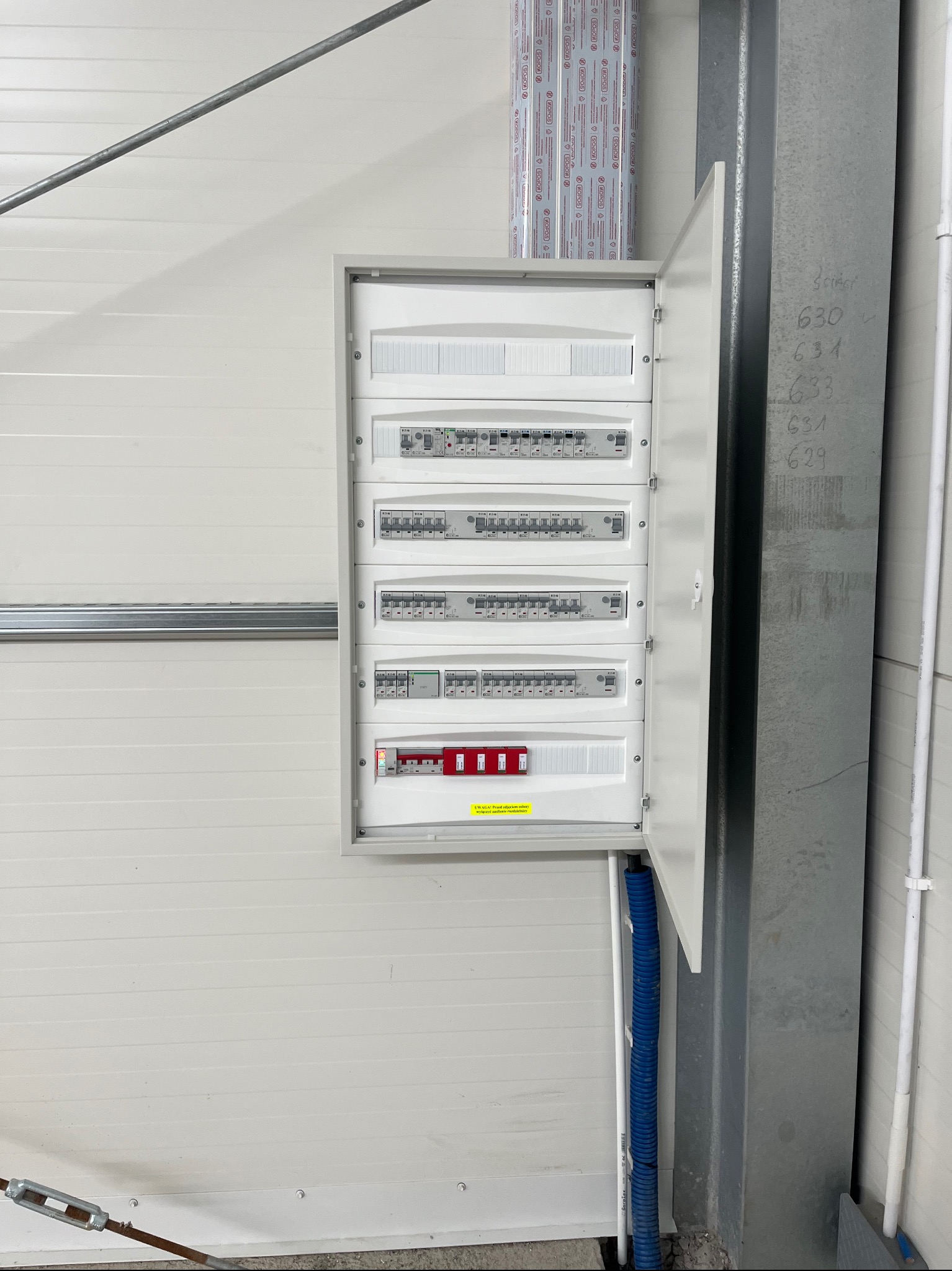 Otwarta skrzynka rozdzielcza z bezpiecznikami i wyłącznikami różnicowoprądowymi zamontowana na białej ścianie z blachy trapezowej, widoczna niebieska rurka karbowana wprowadzająca przewody.