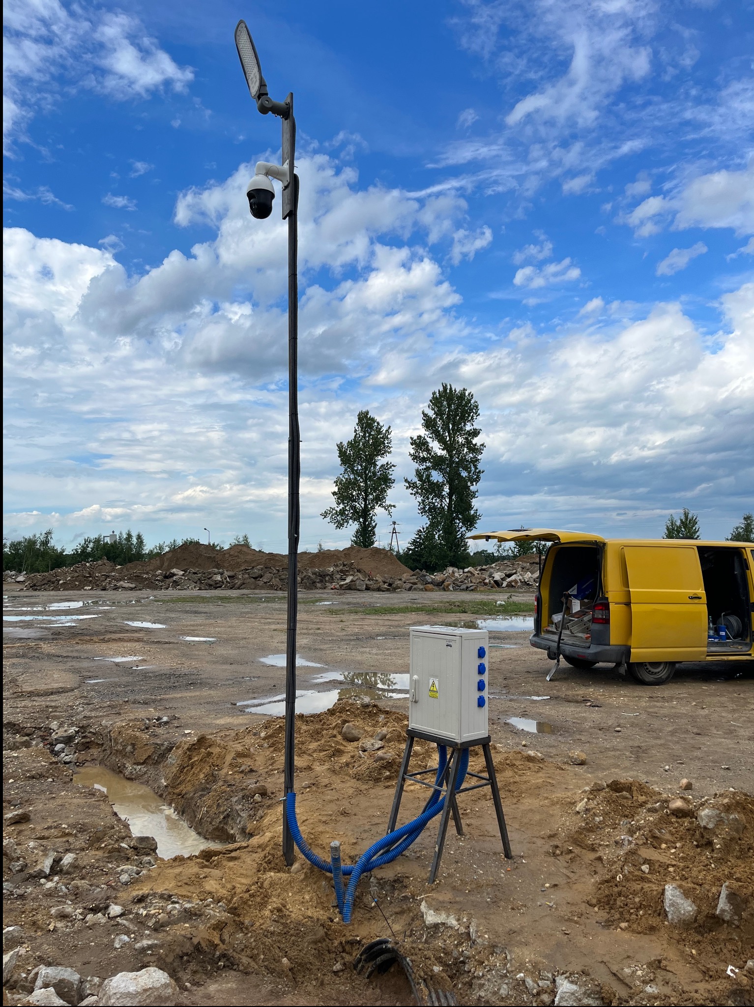 Montaż słupa oświetleniowego z kamerą i podłączeniem elektrycznym na placu budowy, widoczna skrzynka elektryczna na metalowym stojaku, niebieskie rury osłonowe i żółty samochód dostawczy w tle.