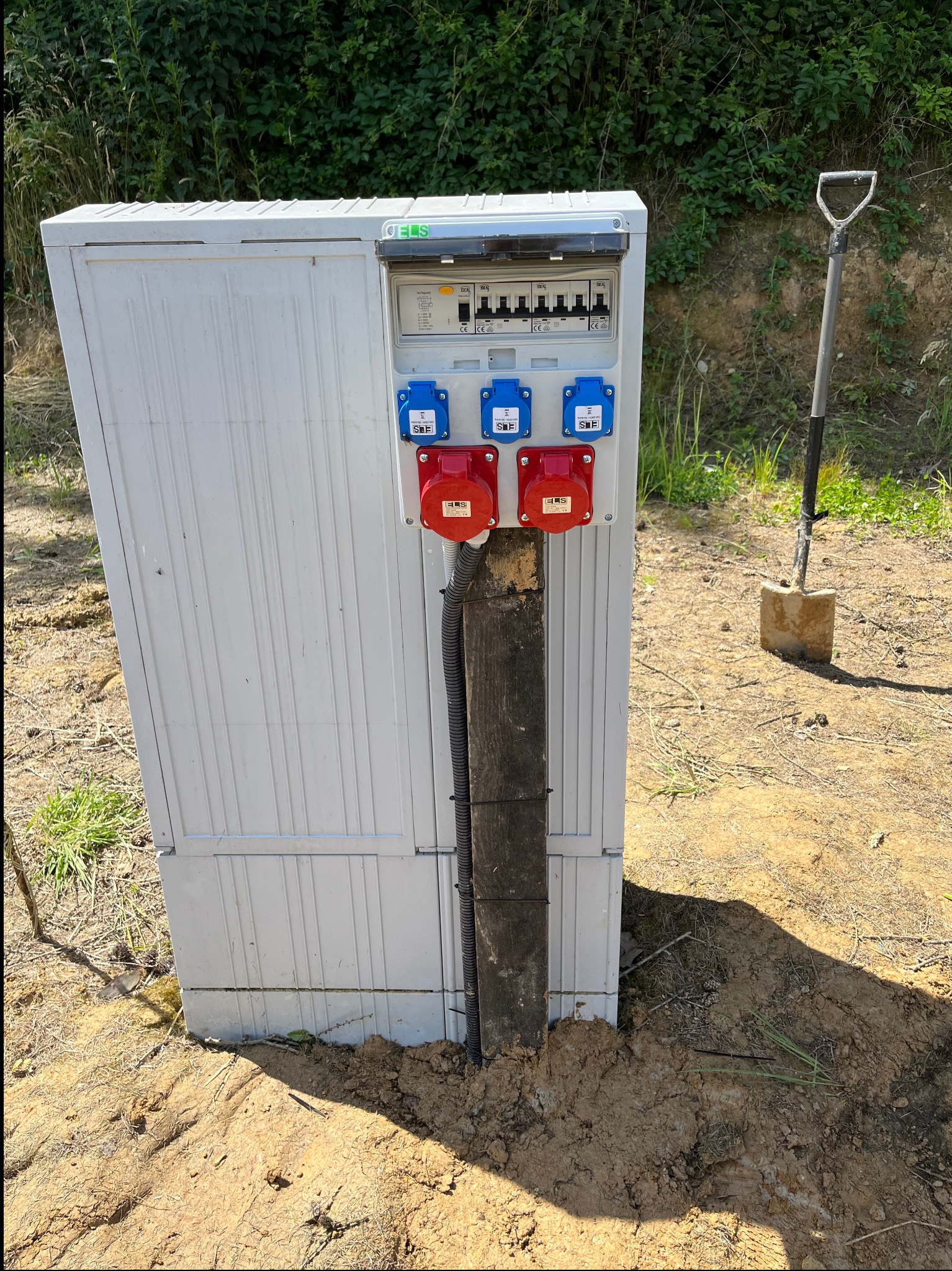 Biała skrzynka elektryczna z gniazdami siłowymi (czerwone i niebieskie) i zabezpieczeniami, umieszczona na drewnianym słupku, obok szpadel wbity w ziemię, na tle zieleni.