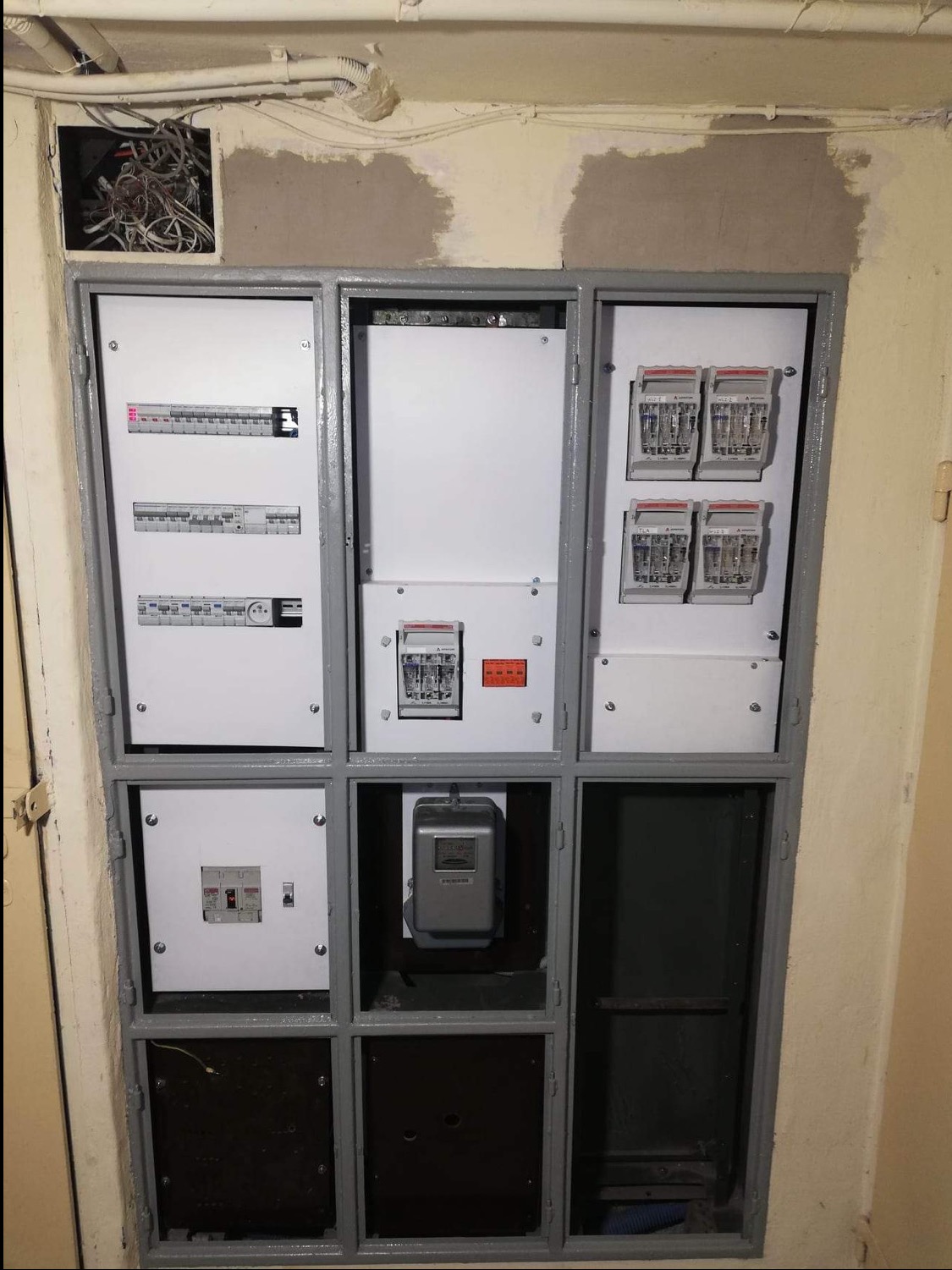 Szafa rozdzielcza z bezpiecznikami, licznikiem energii elektrycznej i przewodami wystającymi z otworu w ścianie, całość po remoncie malowana szarą farbą.