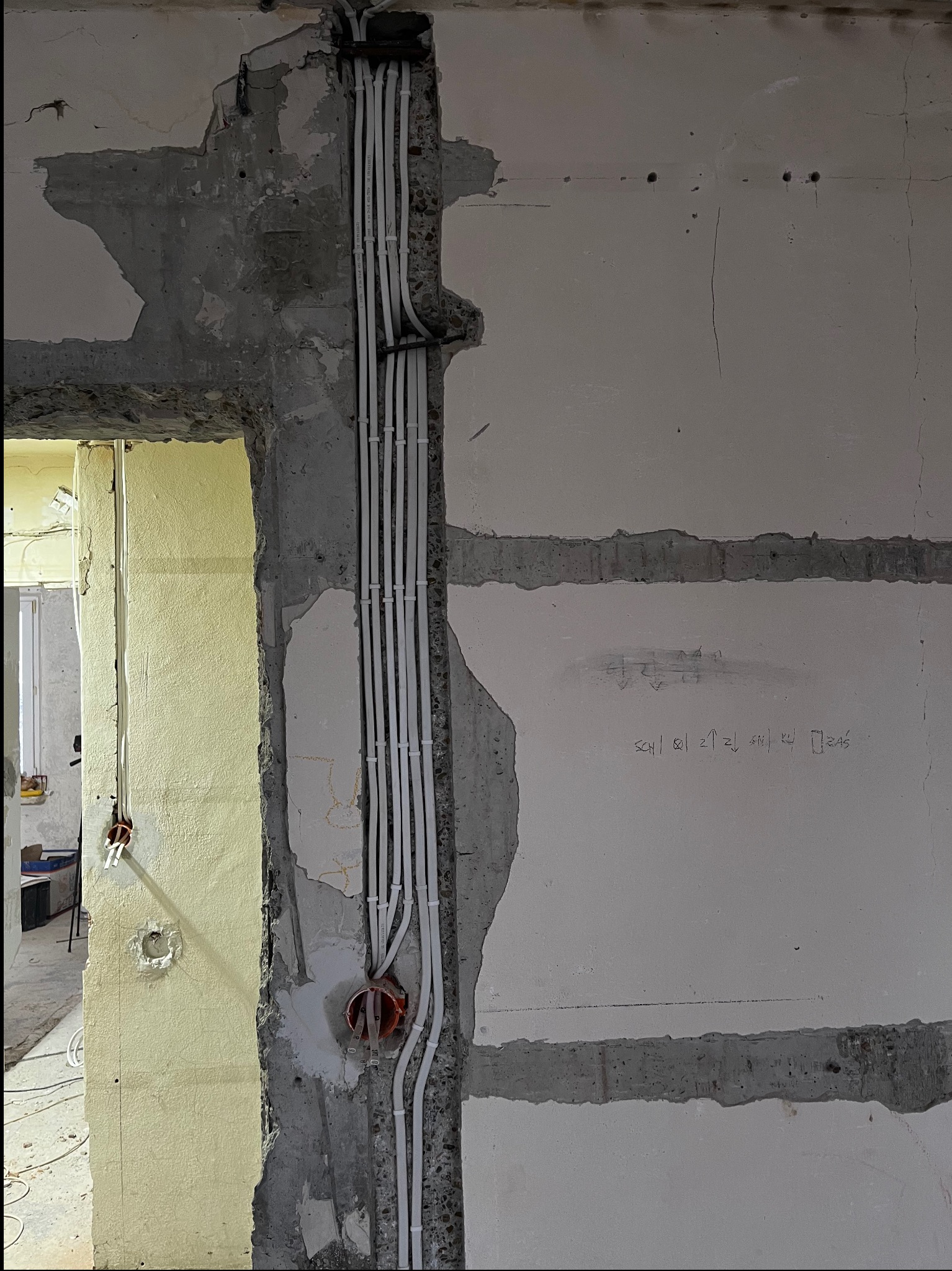 Surowe ściany w trakcie remontu z pionowo ułożonymi białymi rurkami instalacji elektrycznej, widoczne puszki elektryczne, fragmenty starego tynku i betonowa konstrukcja ściany z odręcznymi...