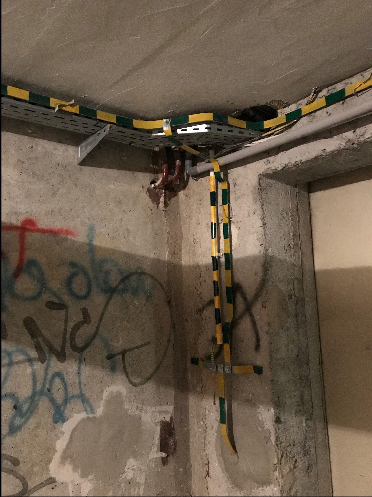 Kable elektryczne w żółto-zielonej izolacji prowadzone po ścianie z graffiti, zamocowane do metalowej drabinki pod sufitem.