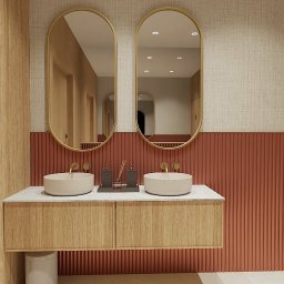 Katarzyna Pepke Zygalska Interiors - Nowoczesna łazienka z dwoma okrągłymi umywalkami nablatowymi, złotymi bateriami i owalnymi lustrami w złotych ramach, na tle ściany z pionowymi, pomarańczowymi panelami i tapety w odcieniach beżu.