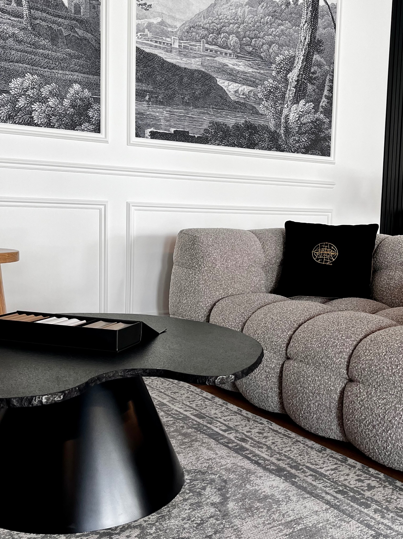 Szary modułowy narożnik z czarną poduszką z geometrycznym złotym wzorem, czarny stolik kawowy o nieregularnym kształcie blatu, dywan w odcieniach szarości oraz czarno-białe grafiki na ścianie...