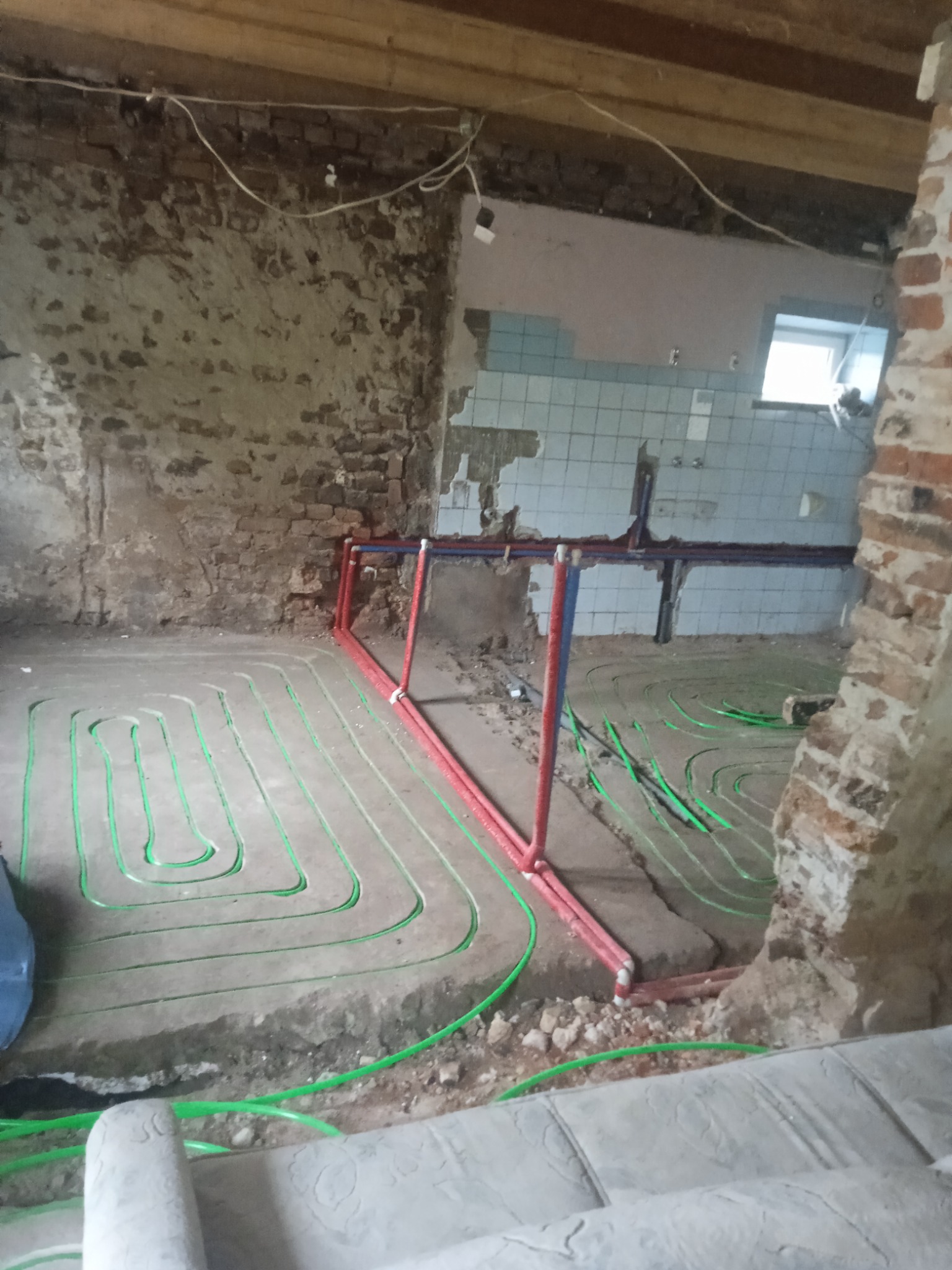 Instalacja ogrzewania podłogowego w trakcie remontu, widoczne zielone rurki w wylewce, fragmenty starej ceglanej ściany i pozostałości płytek na ścianie w tle.