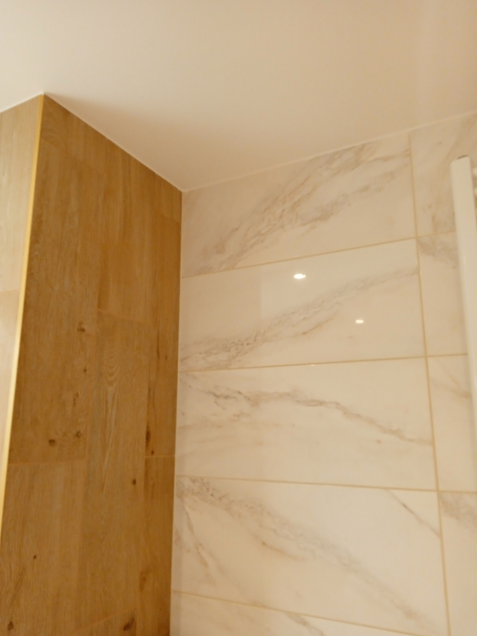 Ściana wykończona płytkami imitującymi marmur w odcieniach beżu, sąsiadująca z pionową powierzchnią wyłożoną płytkami drewnopodobnymi, tworząc eleganckie połączenie materiałów w rogu pomieszczenia.