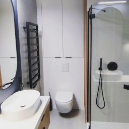 Twoje wymarzone mieszkanie - Nowoczesna łazienka z owalną umywalką nablatową, czarnym grzejnikiem drabinkowym i kabiną prysznicową z czarnymi detalami oraz drewnianymi panelami dekoracyjnymi.