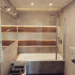 Twoje wymarzone mieszkanie - Nowoczesna łazienka z wanną obudowaną szarymi płytkami, szklanym parawanem nawannowym, drewnopodobnymi akcentami na ścianie i oświetleniem punktowym.