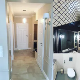 Korytarz z beżowymi ścianami i jasnymi płytkami podłogowymi, widok na białe drzwi i łazienkę z białymi płytkami i toaletą podwieszaną, dekoracyjna mozaika nad lustrem.