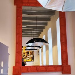 Twoje wymarzone mieszkanie - Odbicie w lustrze w czerwonej ramie: kolumny, betonowy strop, lampy w kształcie parasoli, plakat z żółtą taksówką i osoba robiąca zdjęcie telefonem.