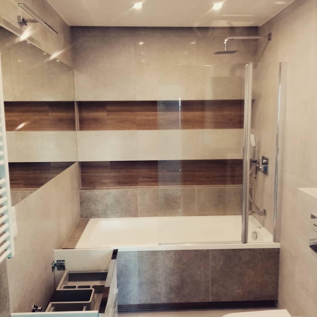 Nowoczesna łazienka z wanną obudowaną szarymi płytkami, szklanym parawanem nawannowym i drewnopodobnymi akcentami na ścianie, szafka pod umywalkę z otwartymi szufladami.