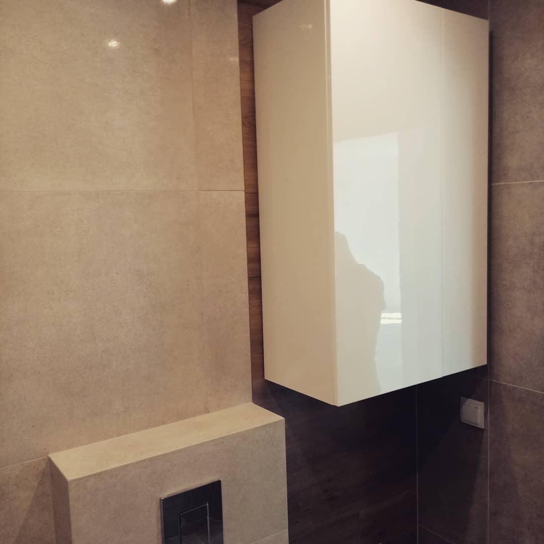 Nowoczesna łazienka z beżowymi płytkami, podwieszana biała szafka z połyskiem i zabudowany system spłukiwania WC z chromowanym przyciskiem.