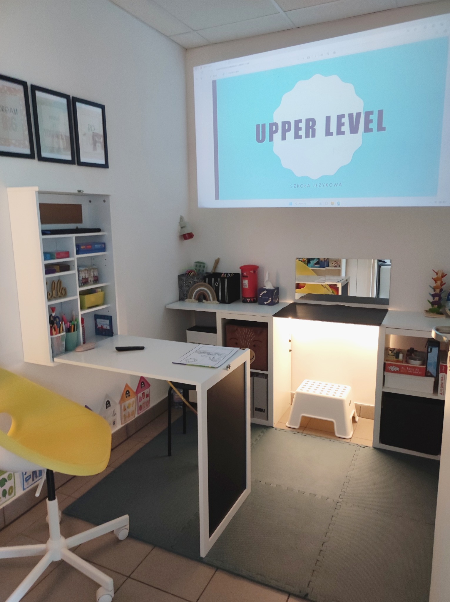 Jasne wnętrze klasy językowej z projektorem wyświetlającym napis 'UPPER LEVEL Szkoła Językowa', białe biurko z rozkładanym blatem, żółte krzesło obrotowe i regały z pomocami dydaktycznymi.