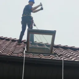 Pracownik w kasku i uprzęży na dachu z brązową dachówką, stojący obok otwartego okna dachowego, pokazuje dwa kciuki w górę.