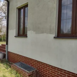 Fragment elewacji domu z dwoma oknami z brązowymi ramami, widoczne ślady niedawnego czyszczenia, ceglana podmurówka, rynna i studzienka rewizyjna przy ścianie.