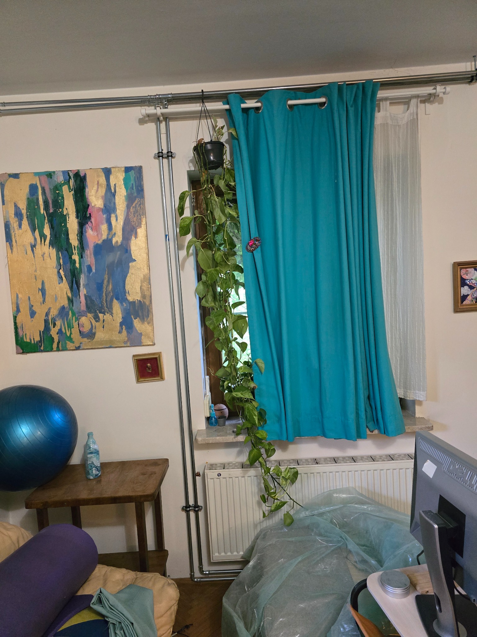 Rury instalacji hydraulicznej poprowadzone na ścianie w pomieszczeniu mieszkalnym, widoczny grzejnik pod oknem, zasłoniętym turkusową zasłoną.