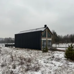 Nowoczesny dom z czarną elewacją i spadzistym dachem na tle zimowego krajobrazu. Widoczny samochód dostawczy i częściowo ogrodzenie. Pochmurne niebo.