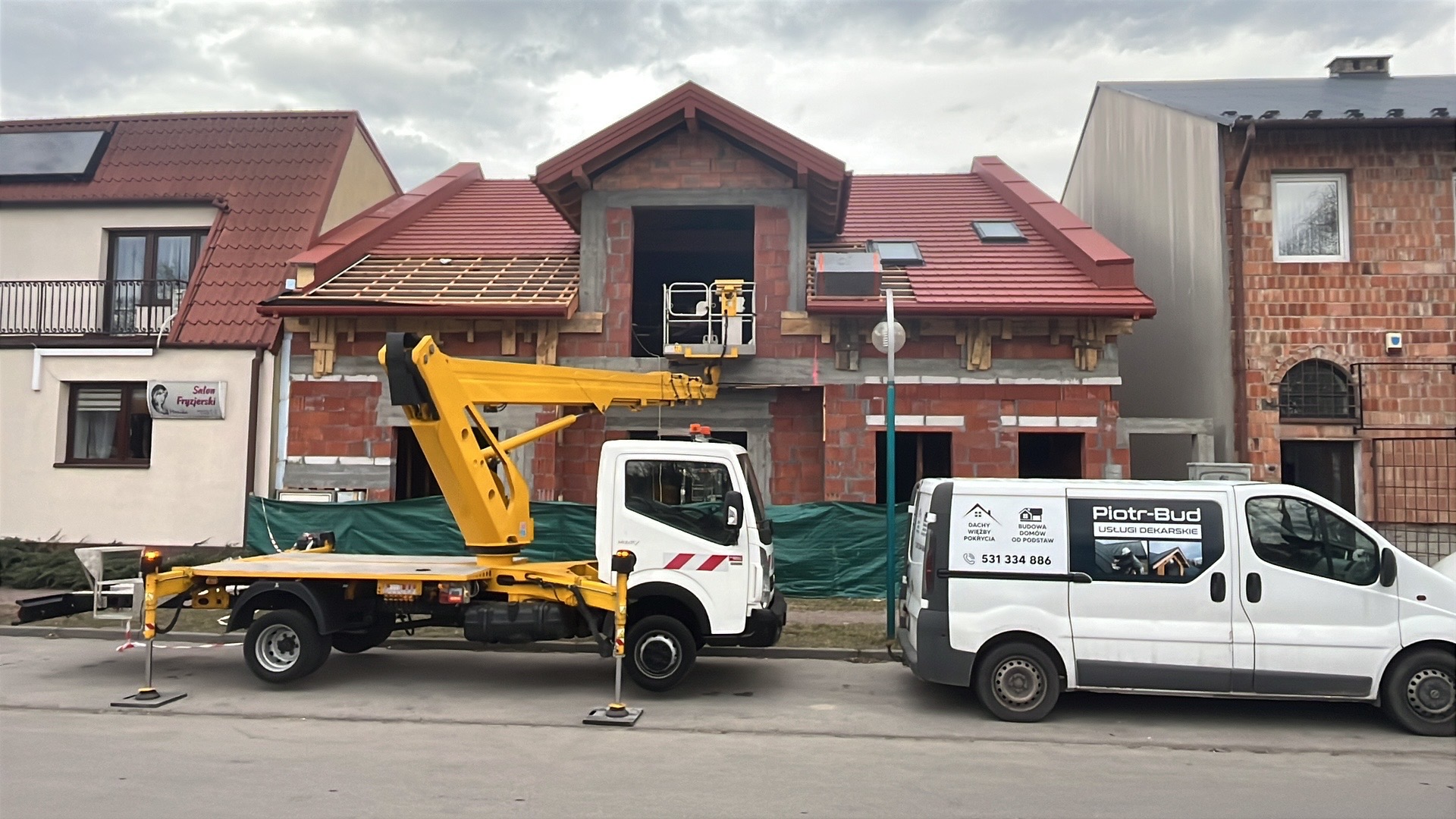 Budowa domu z czerwonej cegły, częściowo pokrytego czerwoną dachówką, z podnośnikiem koszowym i samochodem firmy dekarskiej na pierwszym planie.