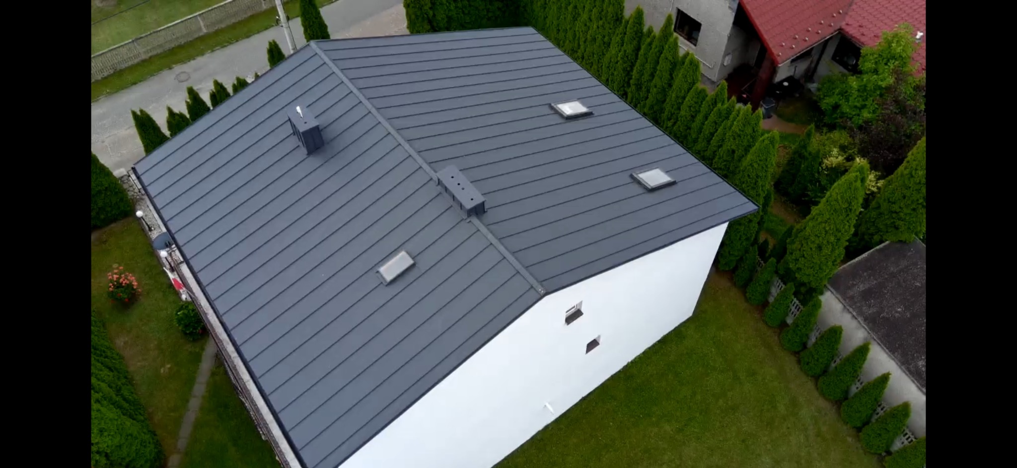 Widok z góry na dom z szarym dachem z blachy na rąbek stojący, z lukarnami i kominem. Biała elewacja, zielony trawnik i tuje. Ujęcie z drona.