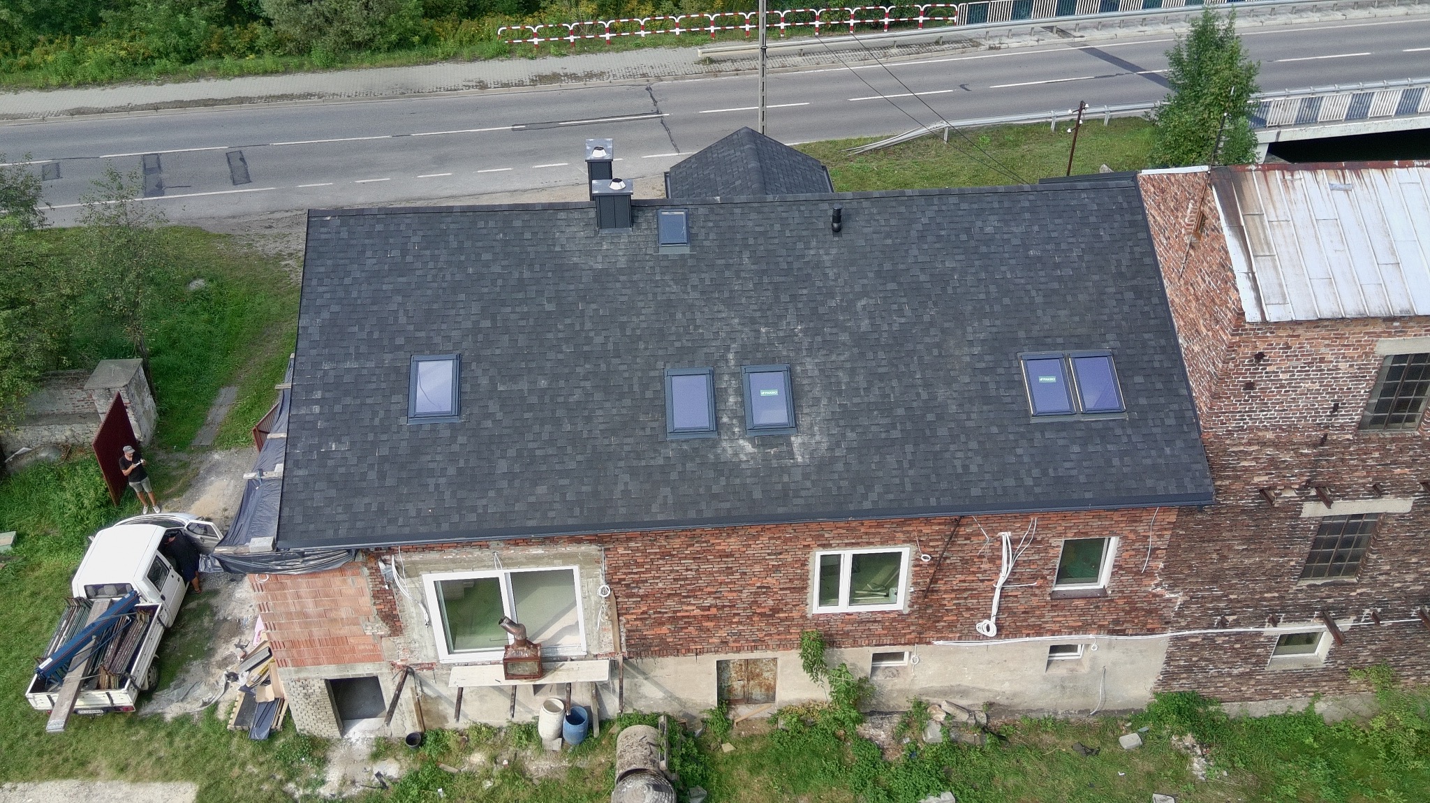Widok z góry na budynek z nowym, ciemnoszarym dachem bitumicznym z oknami dachowymi. Część budynku z cegły, obok zaparkowany biały samochód dostawczy z narzędziami.
