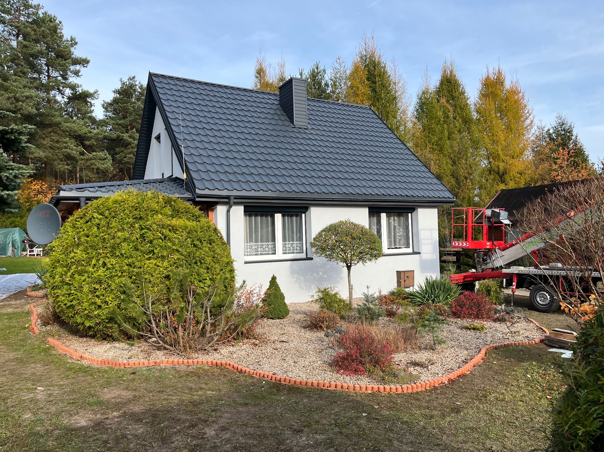 Dom jednorodzinny z ciemnoszarym dachem, otoczony ogrodem. Widoczny podnośnik koszowy, sugerujący prace na wysokości. Jasna elewacja i starannie ułożona dachówka.