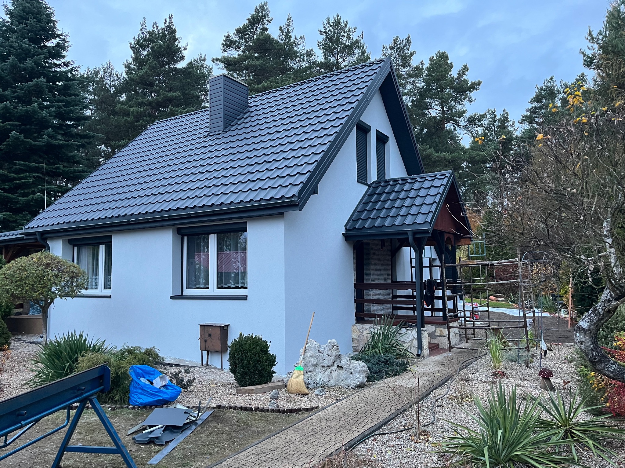Dom jednorodzinny z ciemnoszarym dachem z blachodachówki i kominem. Na pierwszym planie narzędzia blacharskie i fragment ogrodu.