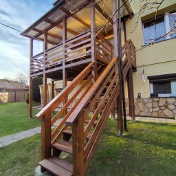 Wojciech Staszel Góral-Drew - Dwupoziomowa, drewniana konstrukcja tarasowa z zadaszeniem, przylegająca do budynku. Schody prowadzą na górny poziom. Widok z dołu, na tle zielonego trawnika i błękitnego nieba.