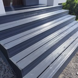 Wojciech Staszel Góral-Drew - Nowoczesne, szare schody wejściowe z desek kompozytowych, prowadzące do drzwi domu. Widoczne detale montażu i faktura materiału. Tło stanowi elewacja i zieleń.