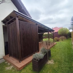 Wojciech Staszel Góral-Drew - Zadaszony taras z drewnianą zabudową, widok z boku. Ciemne drewno, pionowe deski, rośliny w donicach. Tło: trawnik, krzewy, dom z czerwoną dachówką.