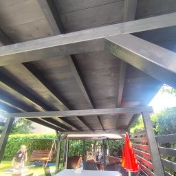 Wojciech Staszel Góral-Drew - Ciemna, drewniana altana z widoczną konstrukcją dachu, stołem nakrytym białym obrusem i czerwonym parasolem. W tle zieleń ogrodu i huśtawka.