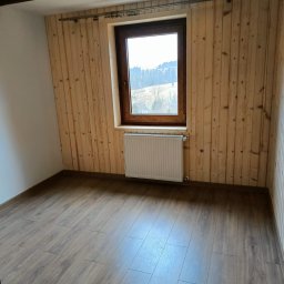 Wojciech Staszel Góral-Drew - Pokój z drewnianą boazerią, brązowymi panelami podłogowymi i oknem z widokiem na zalesione wzgórza. Grzejnik pod oknem. Oświetlenie sufitowe.