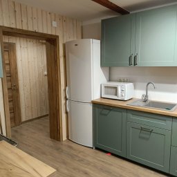 Wojciech Staszel Góral-Drew - Jasna kuchnia z drewnianą boazerią i zielonymi szafkami. Widoczna lodówka, mikrofalówka i zlew. Drewniane belki na suficie dodają rustykalnego charakteru.