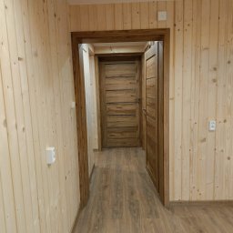 Wojciech Staszel Góral-Drew - Wnętrze wykończone boazerią i drewnianą podłogą. Widoczne dwoje drewnianych drzwi z ciemniejszymi ościeżnicami. Jasne, naturalne oświetlenie.