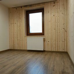 Wojciech Staszel Góral-Drew - Wnętrze pokoju z drewnianą boazerią na ścianach, brązowym oknem i panelami podłogowymi. Widoczny biały grzejnik pod oknem i karnisz. Ujęcie z niskiego kąta.