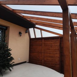 Wojciech Staszel Góral-Drew - Zadaszona konstrukcja z drewna i przezroczystego tworzywa, przylegająca do budynku. Widoczna drewniana ściana i dekoracyjna choinka. Oświetlenie zewnętrzne.