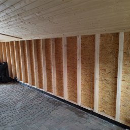 Wojciech Staszel Góral-Drew - Wnętrze w trakcie budowy domu szkieletowego: drewniana konstrukcja ścian z płyt OSB, drewniany sufit, podłoga z kostki brukowej, czarne worki w rogu.