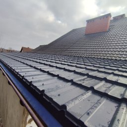 Wojciech Staszel Góral-Drew - Czarny, metalowy dach z ceglanym kominem w pochmurny dzień. Widoczne krople wody na połaci. Ujęcie z dołu, podkreślające strukturę i detale pokrycia dachowego.