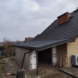 Wojciech Staszel Góral-Drew - Budowa garażu z dachem z dachówki w Suszcu. Widoczne ściany w trakcie wykończenia, ceglany komin i ułożona kostka brukowa. Prace budowlane w toku.