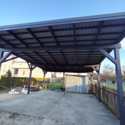 Wojciech Staszel Góral-Drew - Nowoczesna, ciemna wiata garażowa z metalowym dachem i drewnianą konstrukcją, na betonowej wylewce. W tle budynki mieszkalne i zielony ogród.