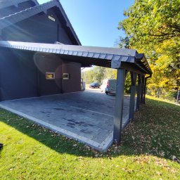 Wojciech Staszel Góral-Drew - Nowoczesna wiata garażowa z ciemną, płaską dachówką, wsparta na solidnych, szarych słupach. Kostka brukowa w odcieniach szarości. Dom w tle, zieleń drzew.