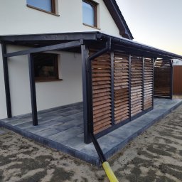 Wojciech Staszel Góral-Drew - Nowoczesna, otwarta konstrukcja z drewnianymi żaluzjami, przylegająca do budynku. Ciemna rama kontrastuje z jasną elewacją i brukowaną podłogą. Widoczny system odprowadzania wody.