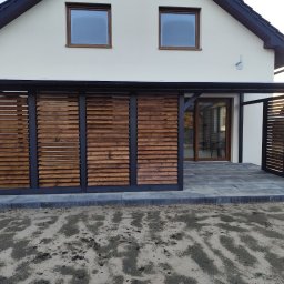 Wojciech Staszel Góral-Drew - Nowoczesna, drewniana zabudowa tarasu z poziomymi lamelami, przylegająca do jasnego domu z ciemnym dachem. Widoczna kostka brukowa i fragment trawnika.