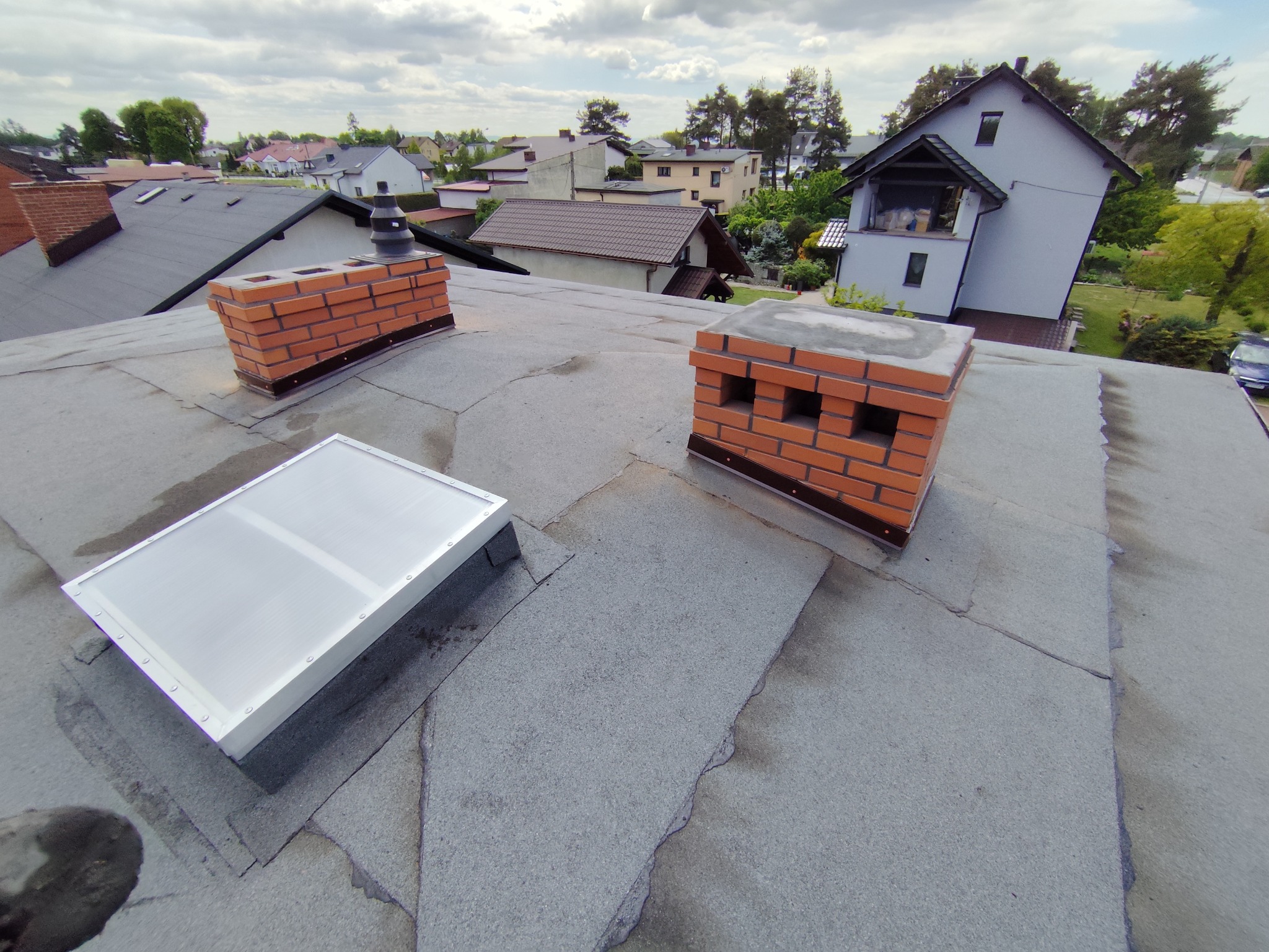Szary dach z papy z dwoma kominami z cegły i świetlikiem. Widok z góry na domy w tle. Pochmurne niebo.