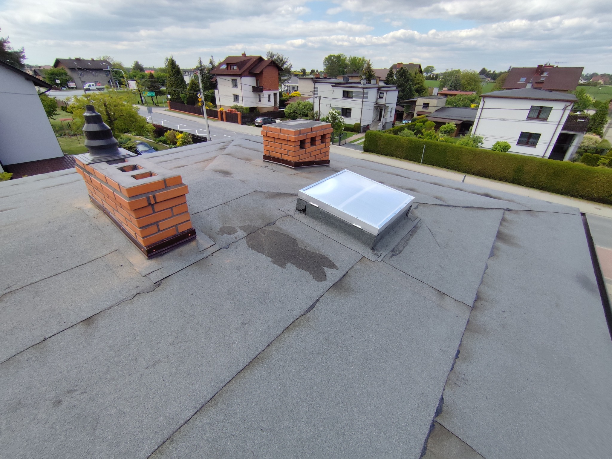 Szary dach z papy z dwoma kominami z czerwonej cegły i świetlikiem. Widok z góry na tle zabudowań i zieleni.