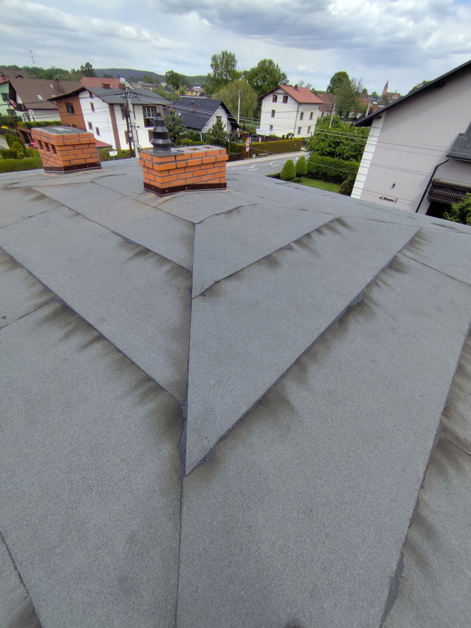 Szary dach z papy z dwoma kominami z cegły na tle zabudowy miejskiej i pochmurnego nieba. Widok z góry, ukazujący detale łączeń papy.