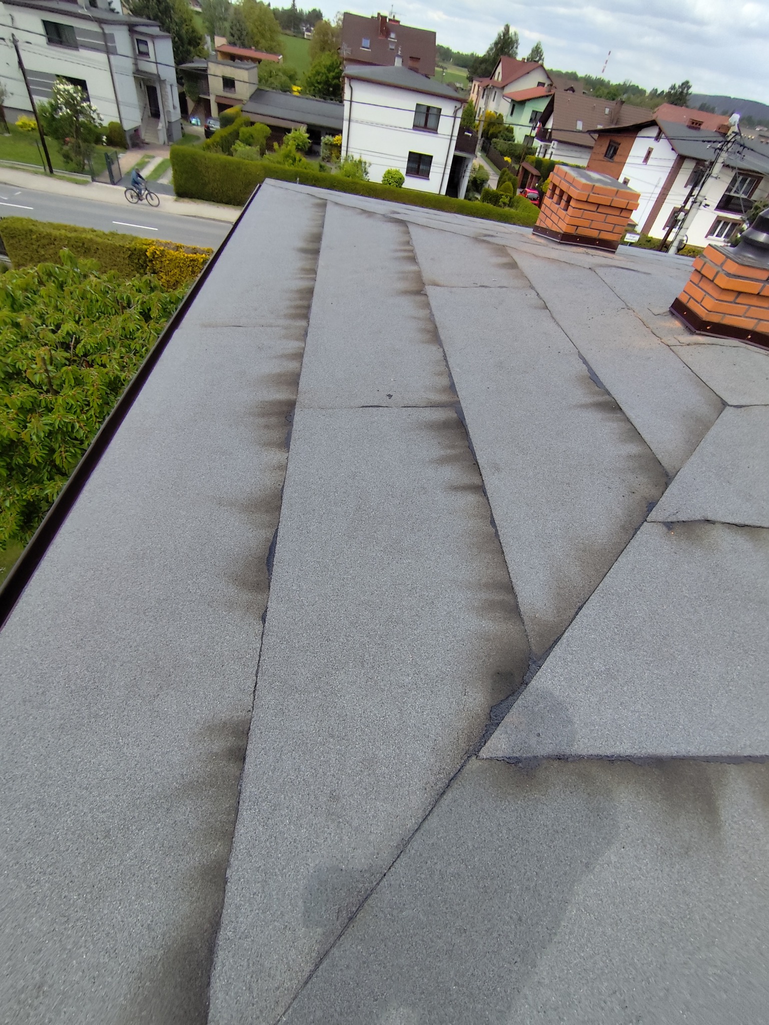 Szary dach pokryty papą termozgrzewalną z widocznymi kominami i otoczeniem domów. Ujęcie z góry, lekko zniekształcone perspektywą.